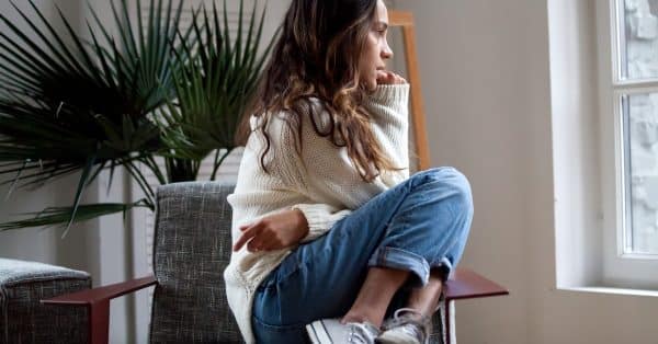 A girl sitting on a lounge chair staring out the window next to her with an anxious look on her face.