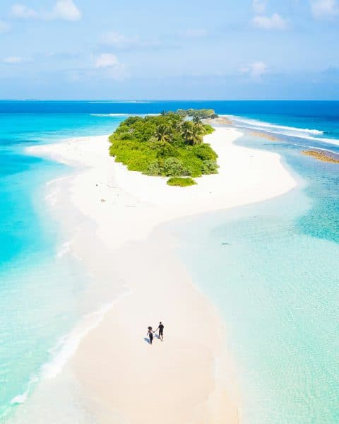 Picture of couple walking along the sand. Surrounded by water, heading to greenery.