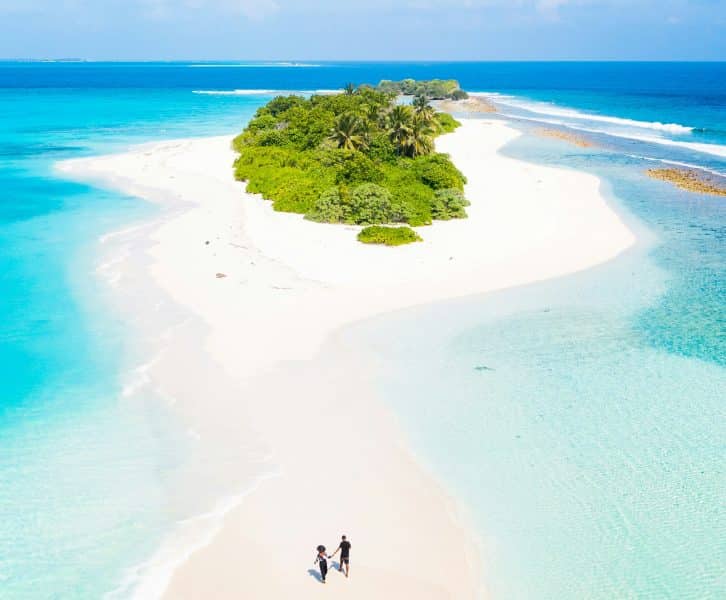 Picture of couple walking along the sand. Surrounded by water, heading to greenery.