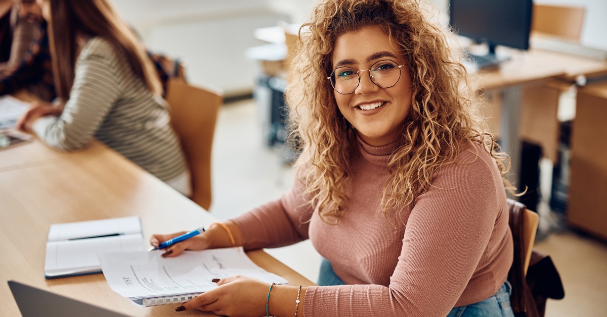 A smiling young woman sits at a wooden table holding a notebook and pen. An open laptop sits in front of her.