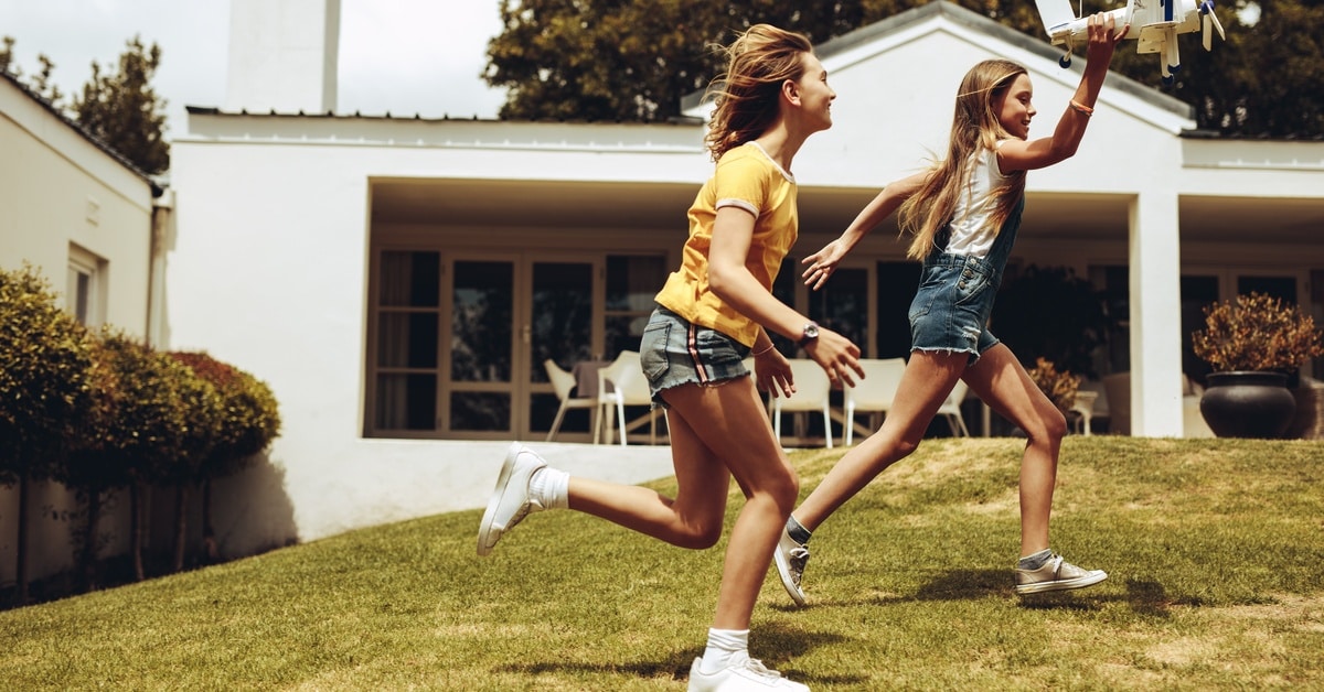 Two girls run across a grassy yard while one holds a toy airplane, with a house and patio in the background.